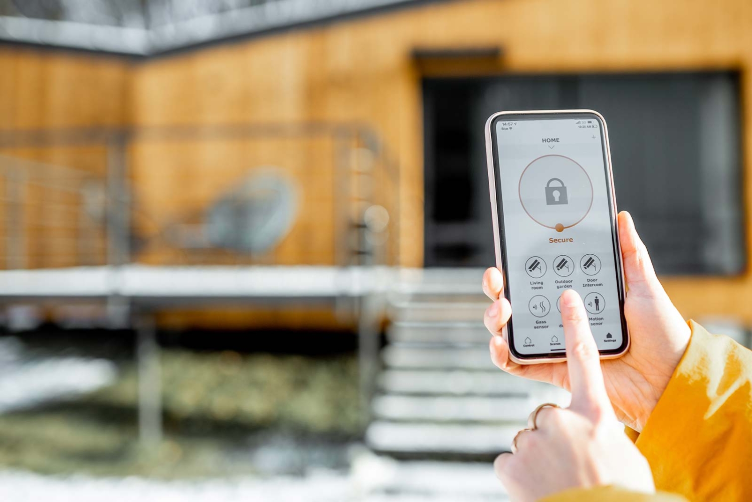 Hands holding smartphone displaying home security app with lock status and various security controls in a modern kitchen